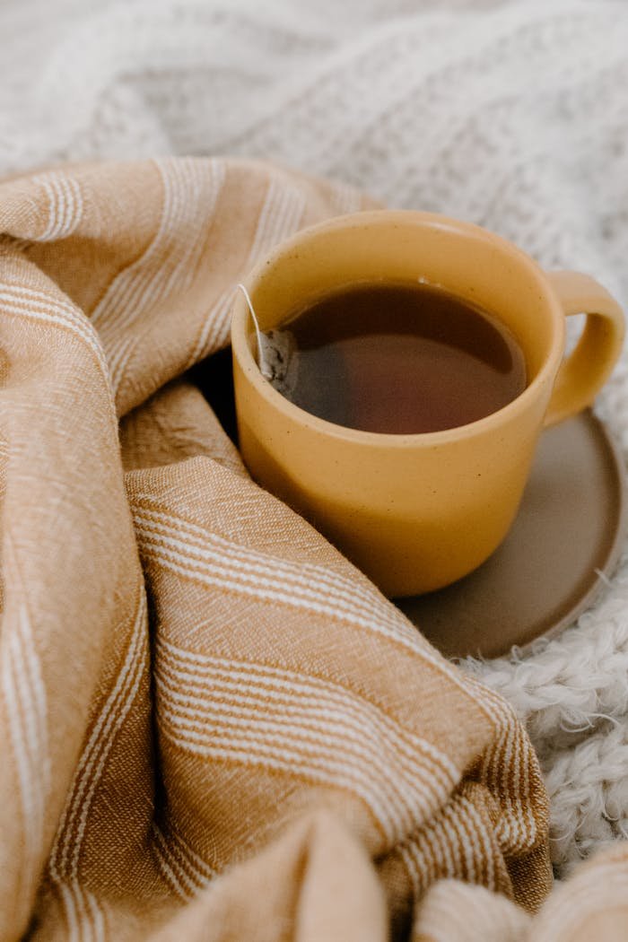 Yellow cup of tea with a teabag nestled in warm, patterned textiles portraying a cozy atmosphere.