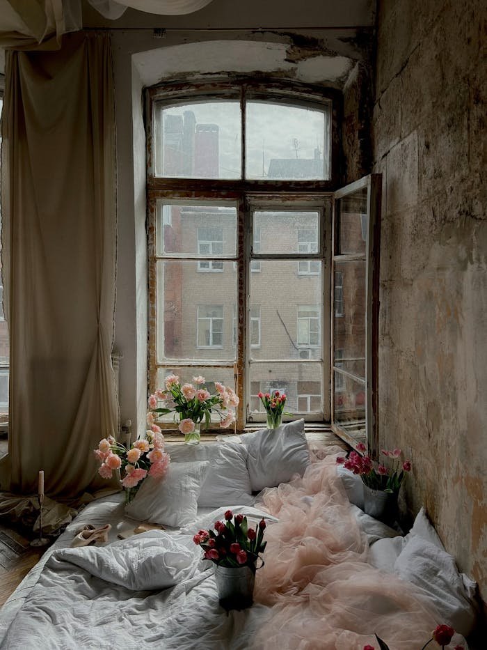 Rustic bedroom with open window, white linens, and colorful flowers creating a romantic vibe.